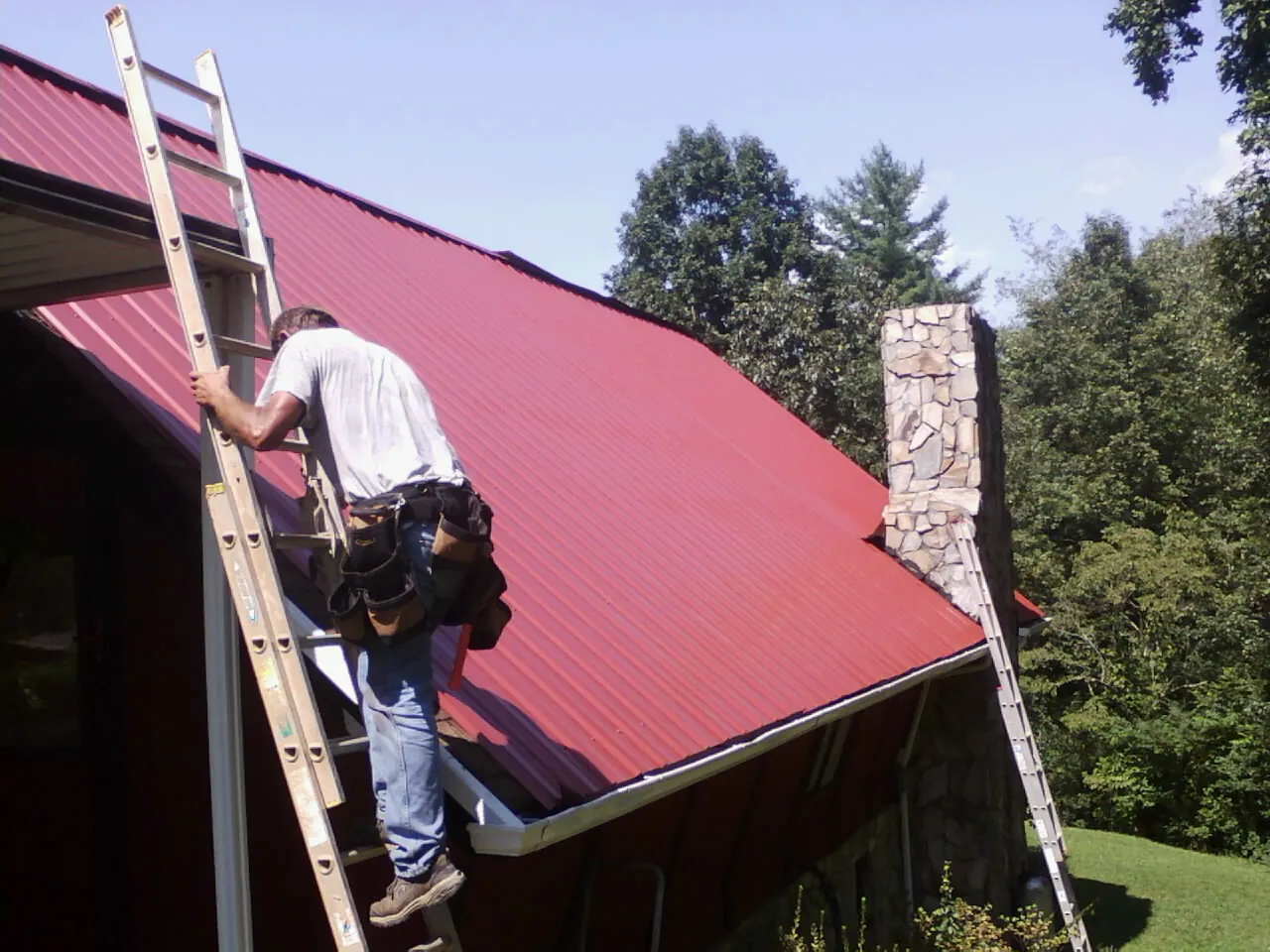 Roofer on a ladder beside a red metal roof and stone chimney — Soffit & Fascia Repair by Roofing Brothers in Blencoe